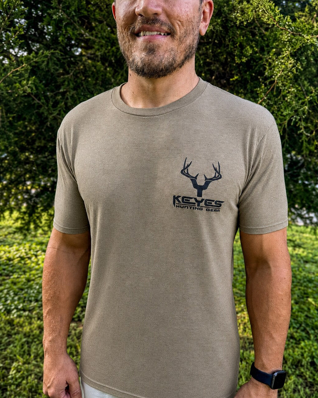 A man wearing a tan short-sleeve t-shirt with a black deer head logo and the text 'KEYES HUNTING GEAR' on the left chest, standing outdoors in front of green foliage.