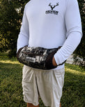 A person outdoors wearing a white long-sleeve shirt and light shorts, with a camouflage-patterned waist bag worn around the waist. The bag has a visible KEYES brand patch and a front zip pocket.
