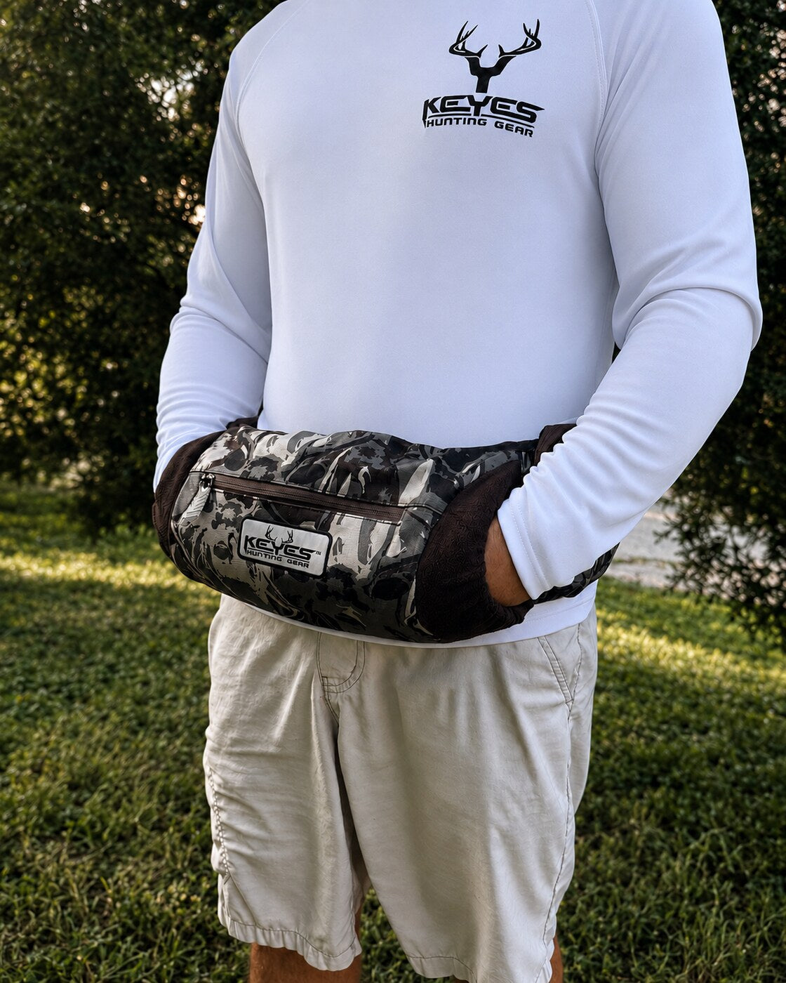 A person outdoors wearing a white long-sleeve shirt and light shorts, with a camouflage-patterned waist bag worn around the waist. The bag has a visible KEYES brand patch and a front zip pocket.