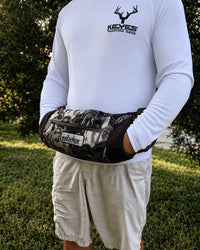 A person outdoors wearing a white long-sleeve shirt and light shorts, with a camouflage-patterned waist bag worn around the waist. The bag has a visible KEYES brand patch and a front zip pocket.