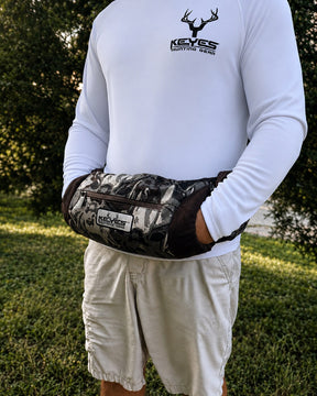 A person outdoors wearing a white long-sleeve shirt and light shorts, with a camouflage-patterned waist bag worn around the waist. The bag has a visible KEYES brand patch and a front zip pocket.
