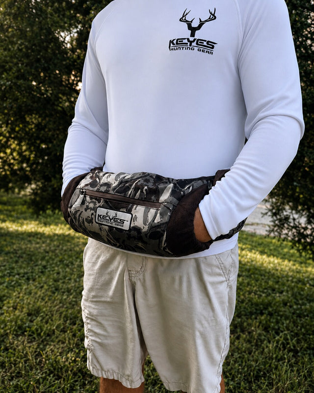 A person outdoors wearing a white long-sleeve shirt and light shorts, with a camouflage-patterned waist bag worn around the waist. The bag has a visible KEYES brand patch and a front zip pocket.