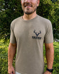A man wearing a tan short-sleeve t-shirt with a black deer head logo and the text 'KEYES HUNTING GEAR' on the left chest, standing outdoors in front of green foliage.