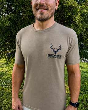 A man wearing a tan short-sleeve t-shirt with a black deer head logo and the text 'KEYES HUNTING GEAR' on the left chest, standing outdoors in front of green foliage.