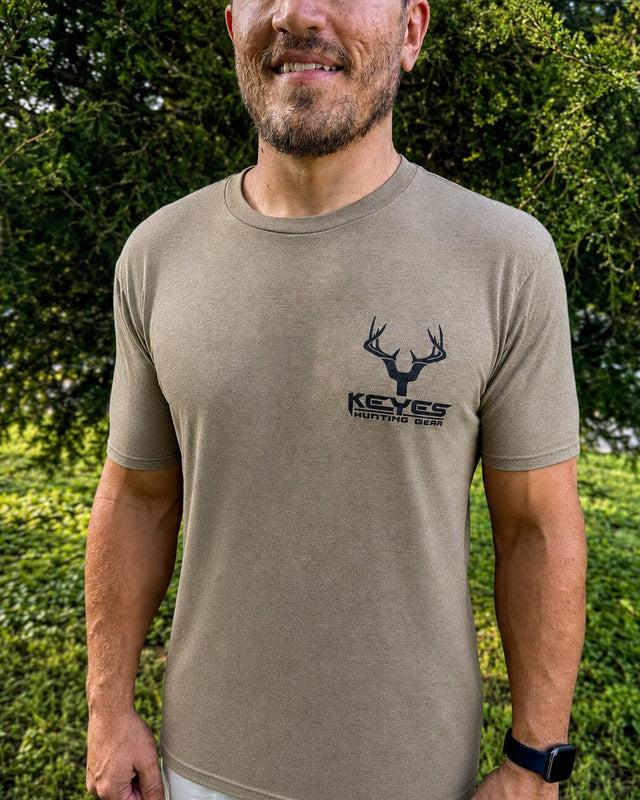 A man wearing a tan short-sleeve t-shirt with a black deer head logo and the text 'KEYES HUNTING GEAR' on the left chest, standing outdoors in front of green foliage.