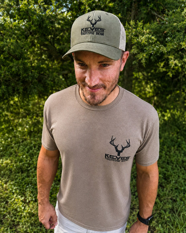 A man wearing a beige short-sleeve t-shirt and a sage green trucker hat, both featuring the 'Keyes Hunting Wear' logo, standing outdoors in front of a green leafy background.